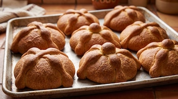 Siete piezas de pan de muerto de masa dorada dispuestas sobre una bandeja metálica de horno.