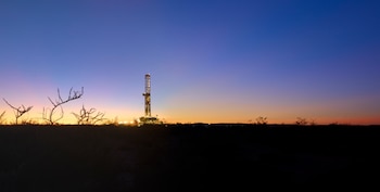 Equipo de perforación de petróleo iluminado, siluetas de vegetación en el primer plano oscuro y un cielo con degradado azul, violeta y naranja