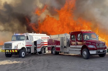 Dos camiones de bomberos, uno blanco y otro rojo, estacionados frente a un gran incendio con llamas naranjas y humo denso