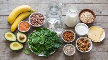 Vista superior de plátanos, aguacate, espinacas, kale, legumbres, frutos secos, avena, lácteos y agua en recipientes sobre una mesa de madera.