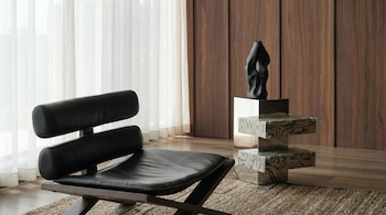 Sillón de cuero negro sobre alfombra, mesa auxiliar de diseño con jarrón escultural frente a paneles de madera y cortinas transparentes.