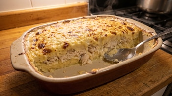 Pastel de papas con pescado gratinado en fuente de horno, con cuchara, vapor visible, sobre tabla de madera y cocina al fondo.