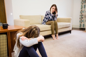 Una niña aparece en primer plano con la cabeza escondida en sus rodillas, sentada en el suelo. Al fondo su madre aparece sentada en el sofá con semblante enfadado
