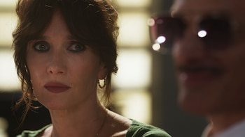 Primer plano de Natalia Oreiro con cabello castaño oscuro, blusa verde y pendientes de aro, mirando al frente. Un hombre con gafas de sol está desenfocado a su derecha