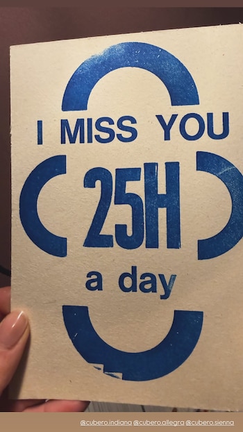 Una mano sostiene una tarjeta beige con un mensaje impreso en azul que dice "I MISS YOU 25H a day", con las letras C y H como arcos