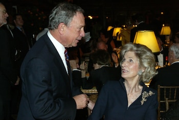 Michael Bloomberg, de traje oscuro, habla de pie con Nancy Kissinger, quien está sentada a una mesa en un salón con lámparas