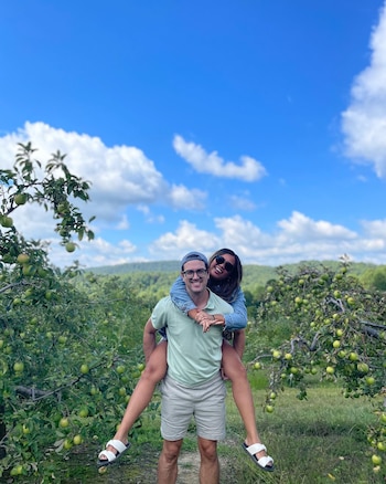 Hombre con gorra y gafas lleva a mujer con gafas de sol sobre su espalda en huerto de manzanos. Árboles con manzanas verdes, cielo azul con nubes blancas
