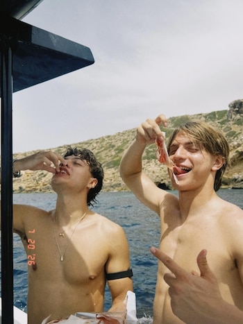 Dos hombres jóvenes sin camiseta comen en un barco, uno de ellos jamón, con el mar azul y una costa rocosa visible al fondo
