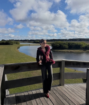 Mujer de pie en muelle de madera, viste chaqueta oscura con cuello y puños peludos, pantalón oscuro, gafas de sol, zapatos rojos, sostiene una copa de vino