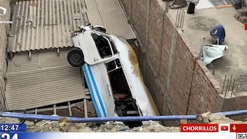 Una combi de color blanco con azul yace inclinada sobre un techo de láminas onduladas entre dos paredes de ladrillos