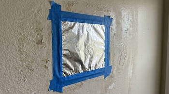 Una pared de color crema con textura y pintura descascarada. Un cuadrado de papel aluminio arrugado está pegado en el centro con cinta adhesiva azul.