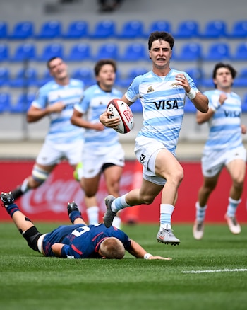 Un jugador de rugby con camiseta celeste y blanca corre con la pelota ovalada. Otro jugador con camiseta azul yace en el suelo en primer plano. Varios jugadores detrás y tribunas azules al fondo