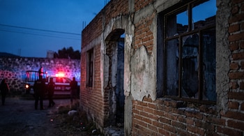 Fachada de ladrillo de una construcción abandonada con ventanas rotas y una patrulla con luces encendidas al fondo al atardecer.