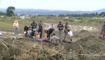 Varias personas, algunas con uniforme de sheriff, trabajan en un terreno de tierra removida con pasto, árboles y colinas al fondo bajo un cielo nublado