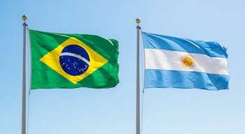 Una bandera de Brasil, verde, amarilla y azul, y una bandera de Argentina, celeste, blanca y con el sol, ondean en mástiles bajo cielo azul.