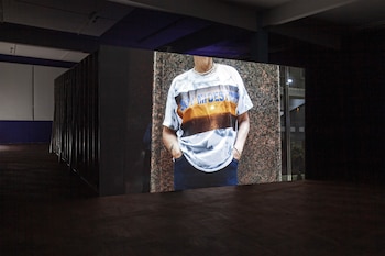 Gran pantalla en una sala oscura muestra el torso de una persona con una camiseta blanca tie-dye que dice 'SOY MI DESTINO' y un atardecer. Manos en bolsillos, suelo de madera