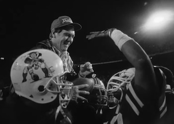 Fotografía en blanco y negro de Raymond Berry sonriendo, con una gorra, sosteniendo un balón de fútbol americano junto a un jugador con casco