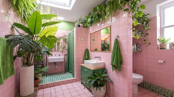 Baño con azulejos rosas y verdes, dos lavamanos, un inodoro, un espejo, toallas verdes, un tragaluz y numerosas plantas en macetas y colgantes.