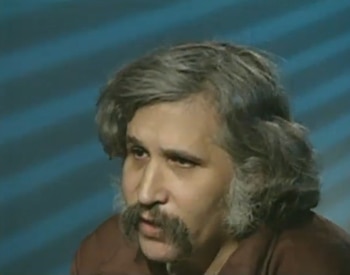 Primer plano de un hombre con cabello gris y bigote poblado, de perfil, mirando hacia la derecha. El fondo es azul con líneas diagonales más claras