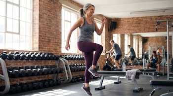 Una mujer madura de cabello gris sonriendo y realizando high knees en un gimnasio. Se ven mancuernas, ventanas y otras personas ejercitándose al fondo.