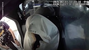 Un oficial de policía con uniforme oscuro y chaleco táctico se inclina junto a la puerta abierta de un vehículo, mientras una persona está parcialmente cubierta en el asiento trasero