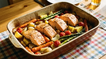 Una bandeja de horno contiene cuatro lomos de salmón rosado, zanahorias anaranjadas, patatas amarillentas, cebollas blancas, tomates cherry rojos y espárragos verdes.