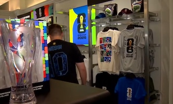 Interior de una tienda de la Copa Mundial 2026. Un hombre de espaldas con camiseta de Messi mira productos. Se ven camisetas, gorras y un trofeo con logo del evento en pantalla