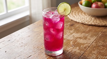 Vaso de cristal con bebida rosa de pitahaya, cubos de hielo, rodaja de limón en el borde, gotas de condensación y mesa de madera.