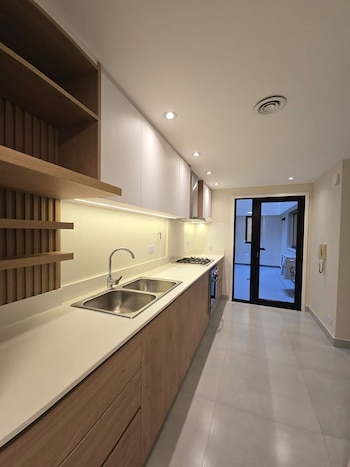 Cocina moderna con gabinetes de madera y blancos, encimera blanca, doble fregadero, estufa, horno y luces empotradas. Puerta de vidrio oscura al fondo