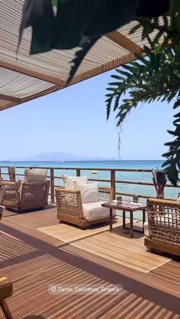 Terraza de madera con techo de pérgola, tres sillones de mimbre con cojines, una mesa, el mar azul, una balsa amarilla y montañas lejanas