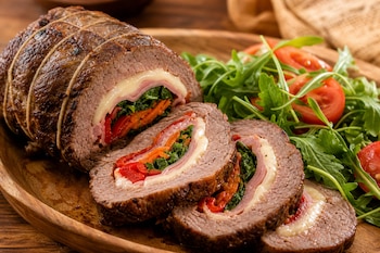 Carne arrollada atada con hilo y cortada en rodajas con relleno de queso, verduras y jamón, servida con ensalada de rúcula y tomate.