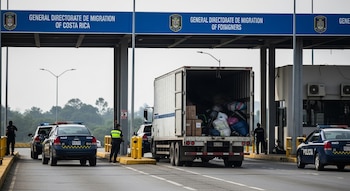 Camión de carga con compartimento abierto, bultos, cajas, tres coches policiales, varios agentes, puesto fronterizo, señalización Dirección General de Migración.
