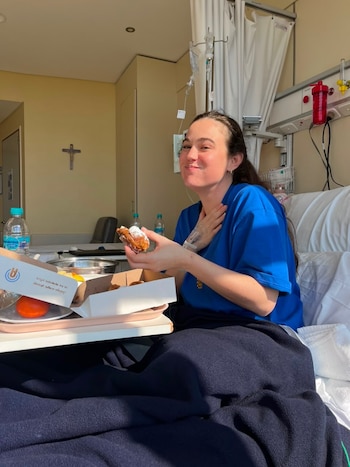 Mujer en cama de hospital con camiseta azul, sostiene un dulce. Bandeja con comida, naranjas y botellas de agua. Equipo médico, cortina y cruz en la pared