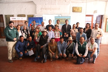 Un grupo de hombres y mujeres posa de pie y agachado frente a banderolas institucionales dentro de una sala con paredes blancas