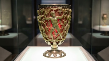 La Copa de Licurgo, un cáliz de vidrio verde oliva con relieves de figuras mitológicas y uvas, iluminado con luz roja que realza sus tallados.