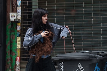 Una mujer de cabello oscuro sujeta una peluca castaña rizada. Al fondo, un contenedor de basura y una persiana metálica cerrada