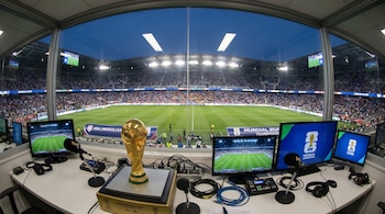 Vista interior de una cabina de transmisión en un estadio de fútbol lleno de gente, con la Copa Mundial de la FIFA dorada en primer plano, monitores y equipos.