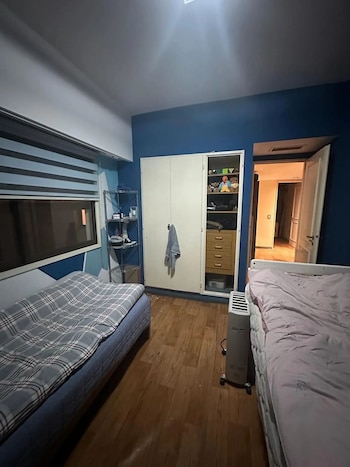 Un dormitorio con paredes azules, dos camas individuales con colchas, un armario blanco y una estantería metálica. Hay una ventana con persianas y suelo de madera