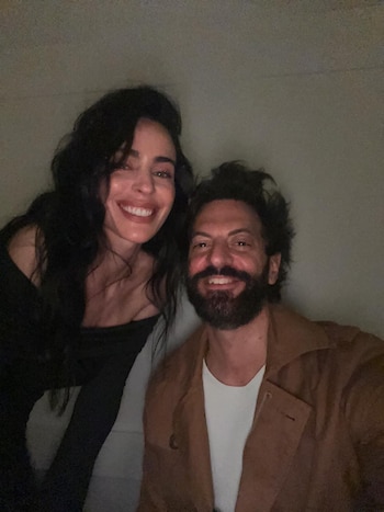Priscila Crivocapich y El Rifle Varela sonriendo a cámara, ella con cabello oscuro y blusa negra, él con barba y chaqueta marrón sobre camiseta blanca