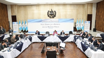 Sala de reuniones con mesa en U y varias personas sentadas con laptops y documentos. Al fondo, banderas de Guatemala y un letrero azul de "Comisión de Postulación" y el escudo
