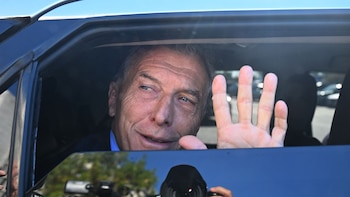 Un hombre de mediana edad, Mauricio Macri, asoma la cabeza por la ventanilla de un coche oscuro y saluda con la mano derecha, bajo un cielo azul