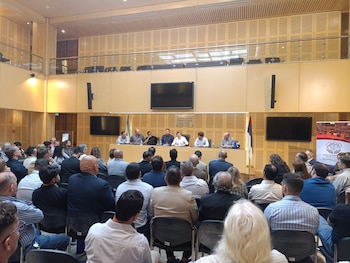 Un panel de siete hombres sentados en una mesa en un salón con paredes de madera y pantallas. Frente a ellos, una audiencia de espaldas. Banderas visibles