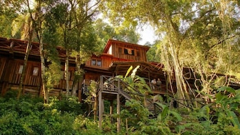Edificio de madera de varios niveles con balcones y escaleras, rodeado de árboles frondosos y vegetación tropical. Dos personas en un balcón