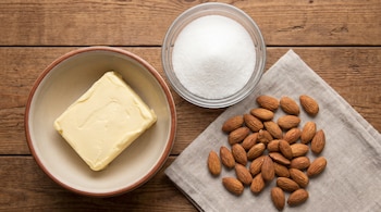 Un cuenco con manteca, un bol de vidrio con azúcar blanca y almendras enteras sobre un paño de tela en una mesa de madera.