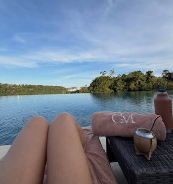 Piernas de una persona recostada en un sillón junto a una piscina de borde infinito, con las Cataratas del Iguazú de fondo, un mate y un termo