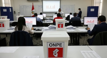 Sala de cómputo con personal de espaldas frente a monitores con gráficos de barras, revisando papeles. Urnas blancas con logo ONPE en primer plano, bandera de Perú.