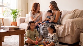 Dos mujeres y tres niños con rostros difuminados, sentados en un sofá y el suelo de una sala con mesa de madera y planta.