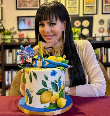 Maribel Guardia, mujer de cabello oscuro y largo, sonríe detrás de un pastel blanco con diseño de limones y flores, coronado por una figura de mujer en traje de carnaval