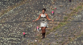 Mujer con vestido brillante y botas camina sobre un cable tenso sosteniendo una barra larga sobre el fondo de una ladera rocosa