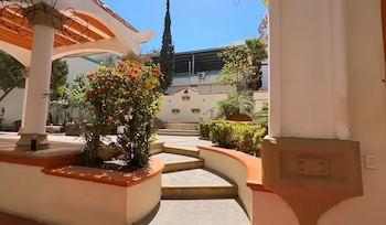 Patio exterior con jardineras, plantas con flores rojas, escalones de piedra y columnas blancas bajo un cielo despejado